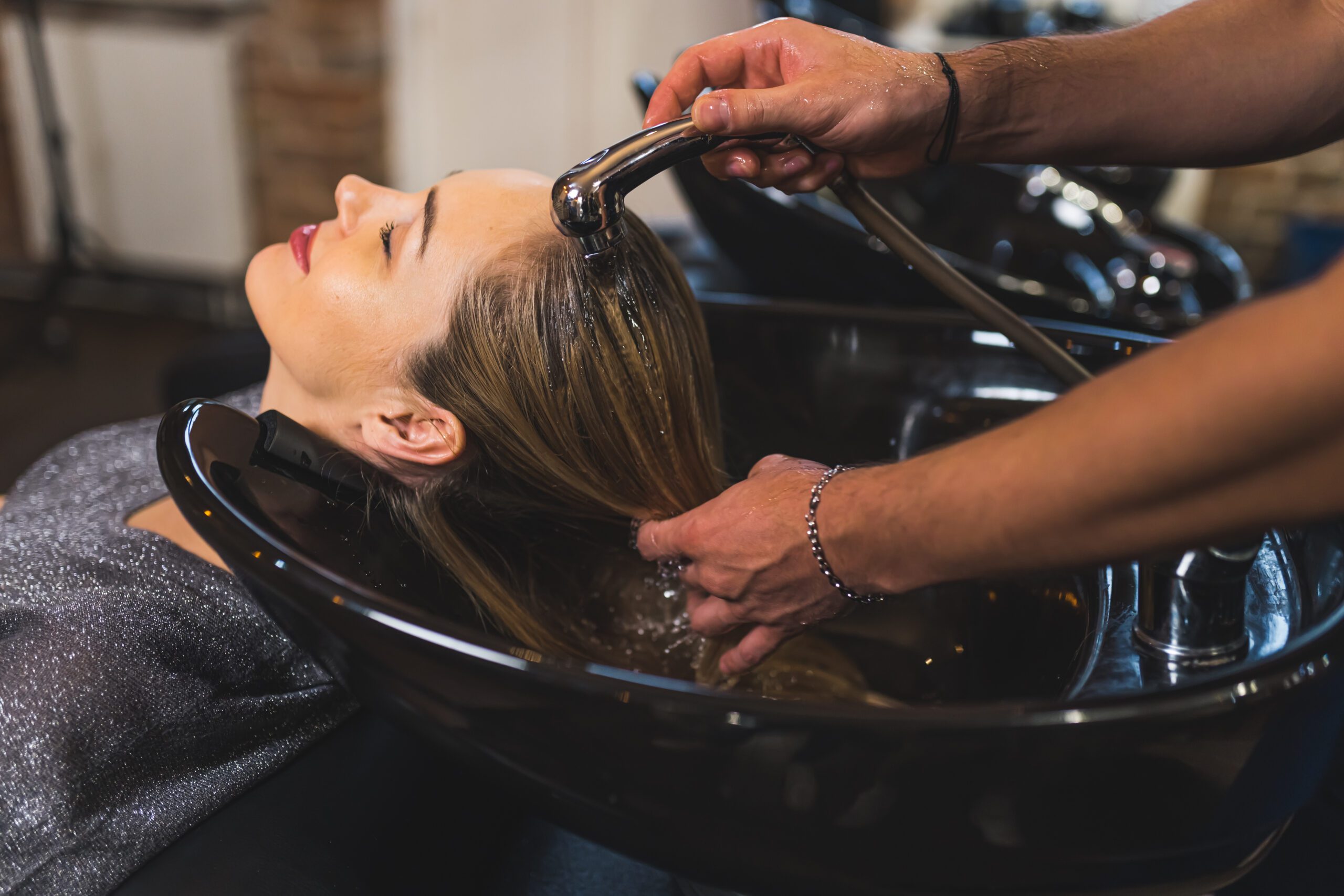 hairdresser washing client’s hair at the salon, side view medium closeup, beauty concept A woman reclining at a salon sink with her head being gently washed by a haircare stylist using a handheld spray nozzle. The setting is a professional hair salon with a relaxing ambiance, and the woman appears to be enjoying the experience.
