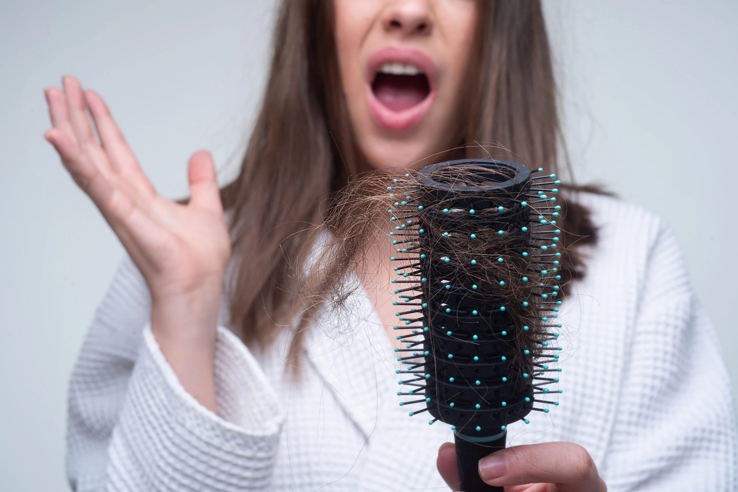 A woman in a white robe holding a round hairbrush tangled with damaged hair, with a shocked and frustrated expression on her face of how to repair her damaged hair. The background is plain and minimalistic.